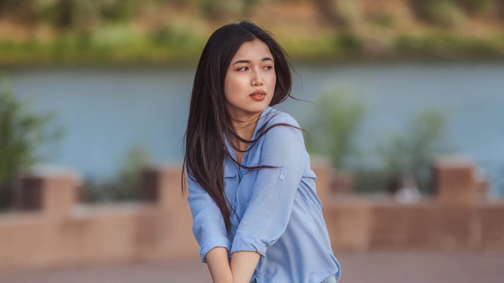 Young woman in a blue shirt posing pensively by the riverside, in a serene, relaxed outdoor setting.